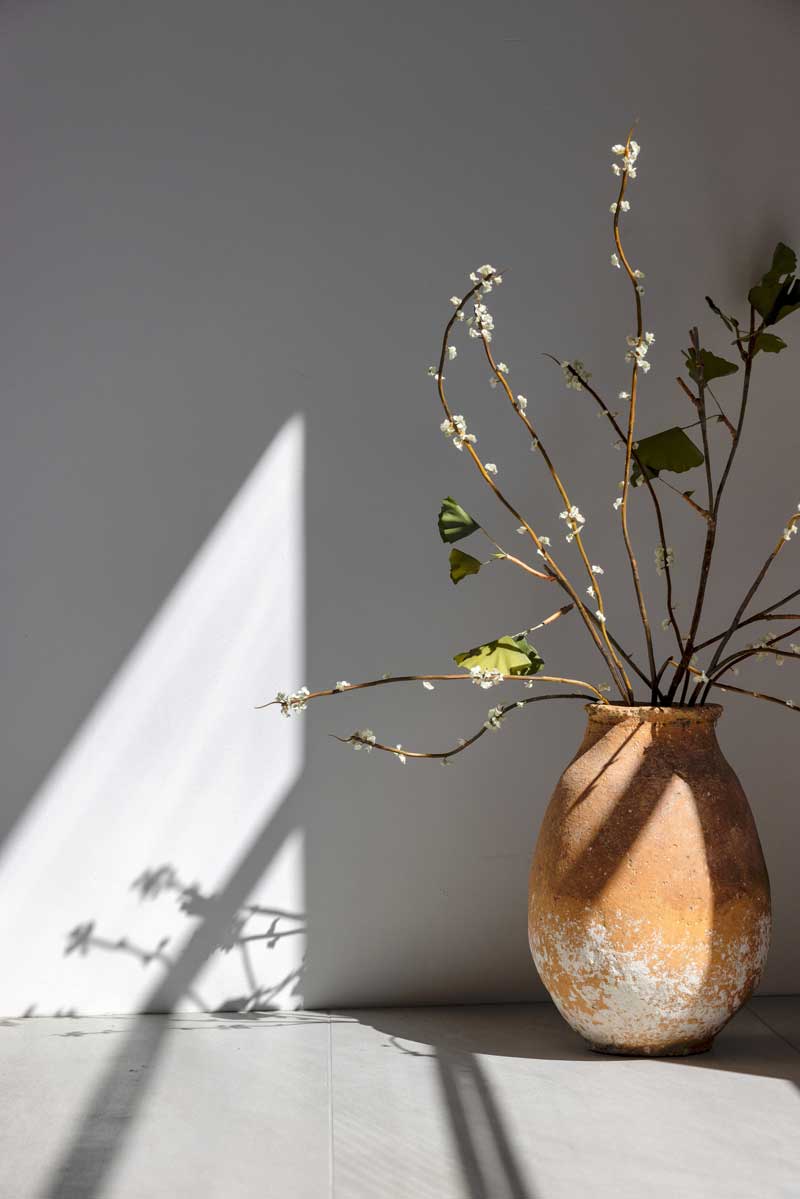 A terra cotta colored vase with branches of flowers on a gray tile floor. A shadow reflects from a Marvin Ultimate window on the wall.