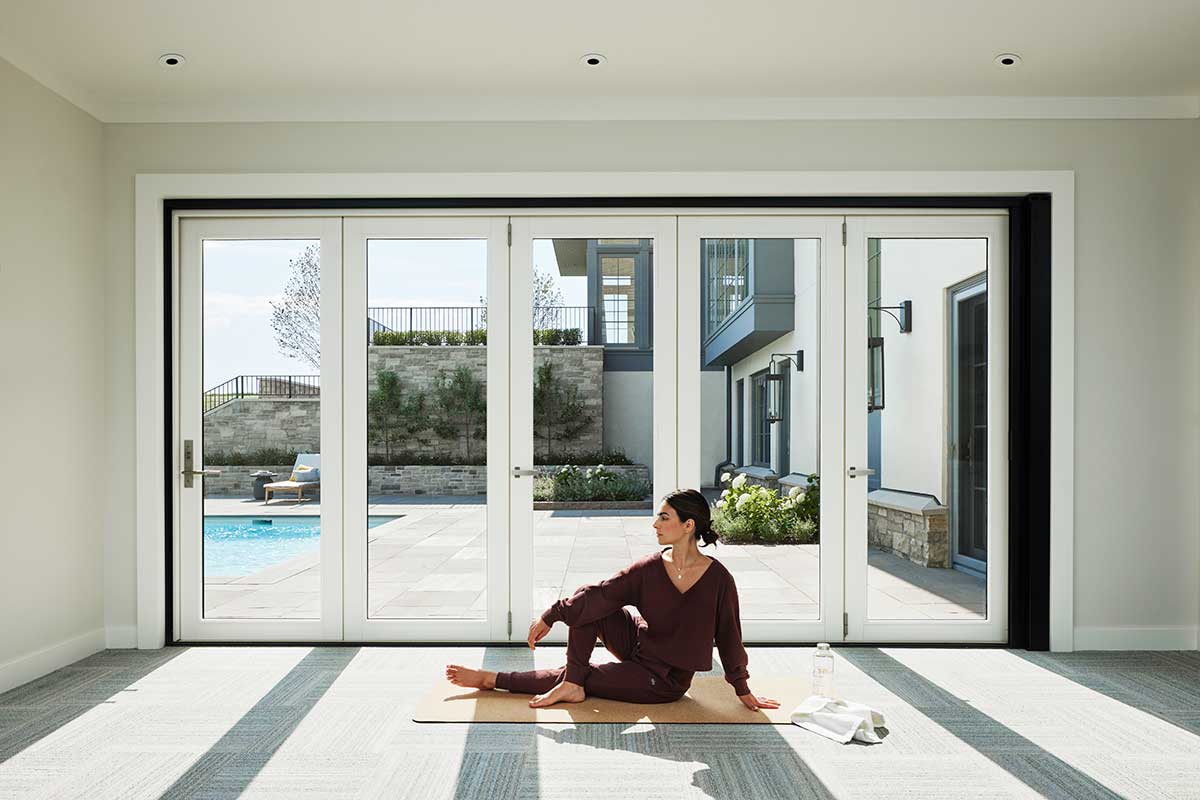 A woman sitting on a yoga mat next to a Marvin Ultimate Bi-Fold door, which looks out to a pool deck and patio.