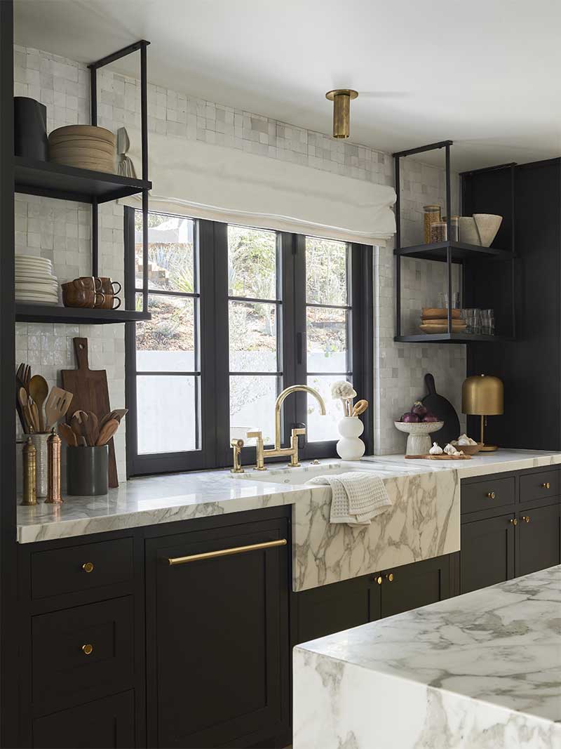 The kitchen at Bobby Berk’s office space, featuring a custom Marvin Ultimate Bi-Fold window, white and gray marble countertops, dark cabinets and open shelves.