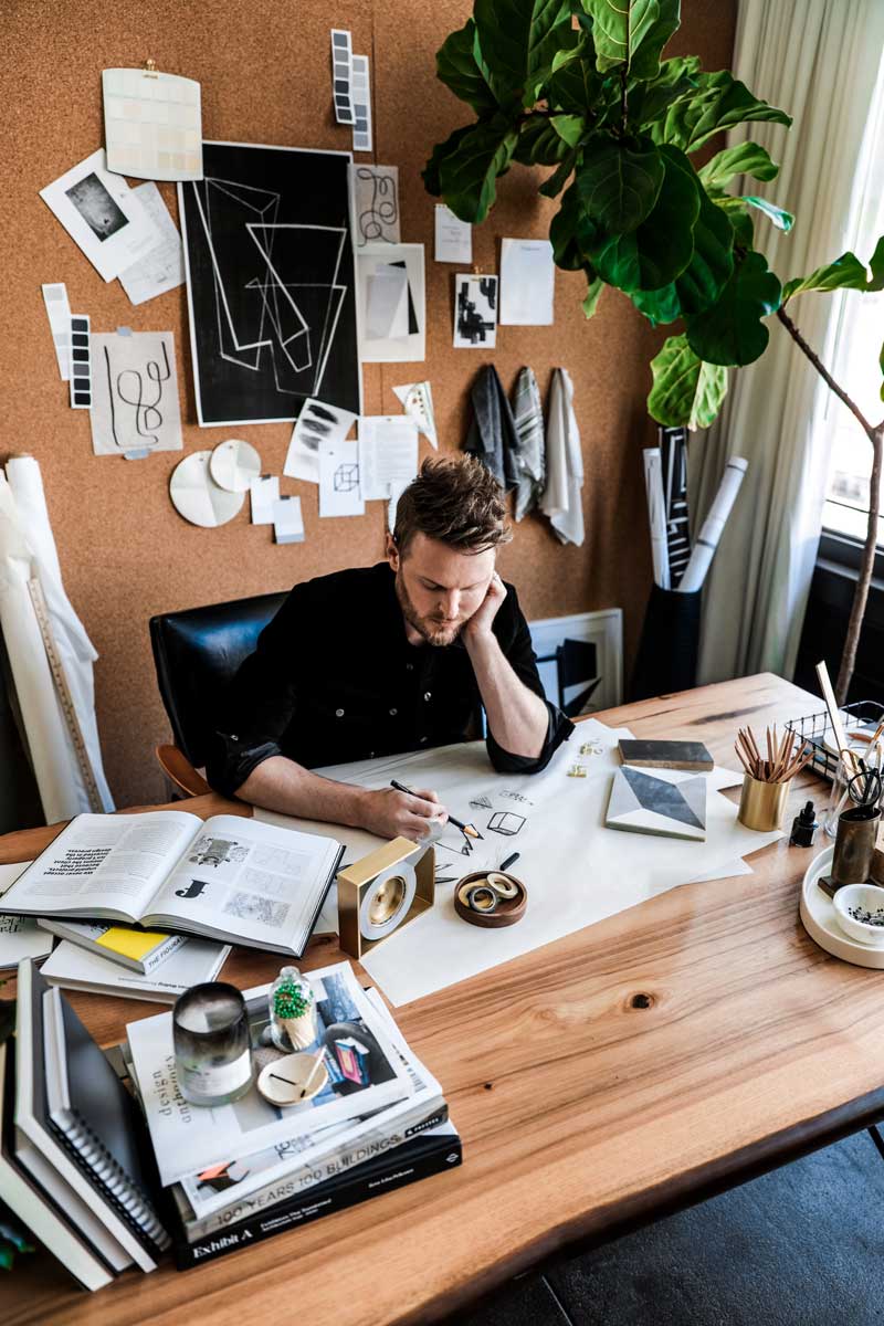 Bobby Berk drawing at his office desk, which is covered with samples, books and other inspiration for his design process.