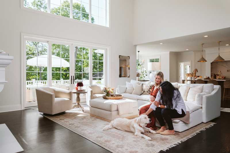 A woman standing behind a couch and a woman petting a dog in a two-story living room featuring Marvin Essential windows.