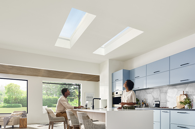 A man and woman in their kitchen, looking up at two Marvin Awaken Skylights with two Marvin Skycoves in the background.