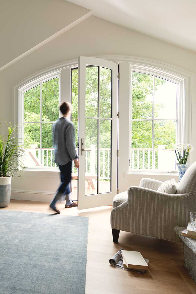 A woman walking through a Marvin Ultimate Archtop French door out to a porch from a bedroom of Hadley House in Wenham, Massachusetts.