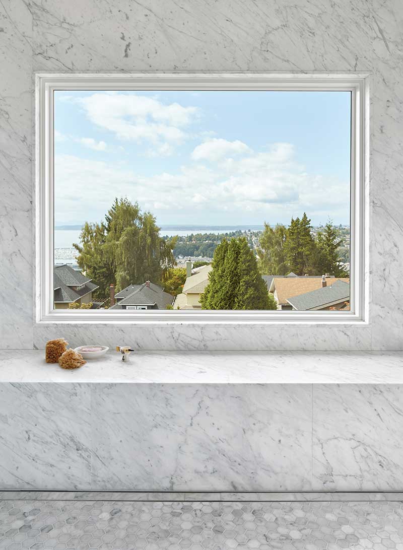 View of rooftops and trees through a Marvin Ultimate Picture window in a contemporary bathroom with marble.
