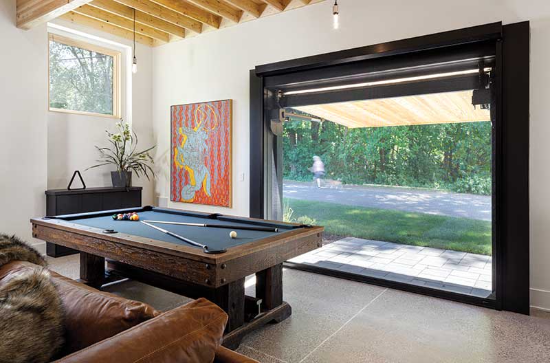 Hydraulic commercial garage door with cedar opens in a modern industrial-style home to reveal a game room with pool table. Marvin Ultimate Picture window is featured. 