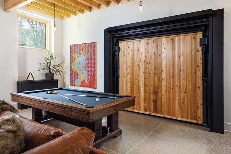 Hydraulic commercial garage door with cedar opens in a modern industrial-style home to reveal a game room with pool table. Marvin Ultimate Picture window is featured. 