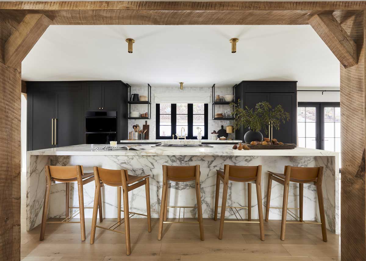 The kitchen inside the Bobby Berk Firm’s office, featuring a custom Marvin Ultimate Bi-Fold window above the sink and a large island with white and gray marble countertops and stools made of wood and leather.