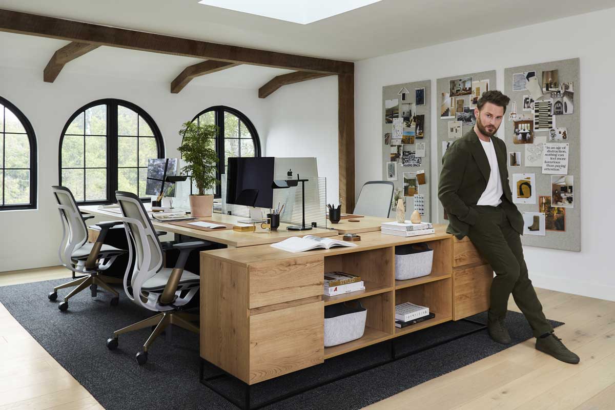 Bobby Berk leaning on a bookshelf in his renovated office space, featuring Marvin Ultimate Round Top windows and three desks with Mac computers on them and mood boards on the wall.