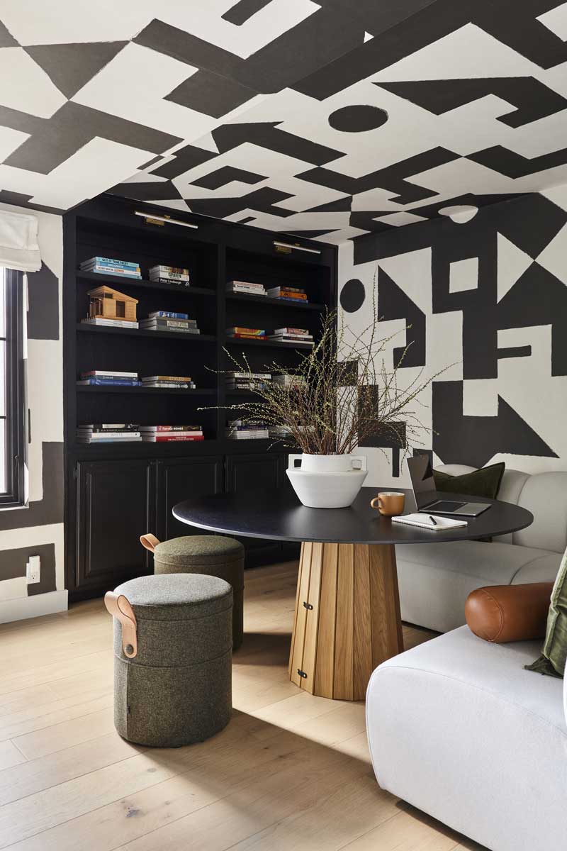 A room in the Bobby Berk Firm’s office with a modern table, banquette and stools and black and white geometric wallpaper on the walls and ceiling.