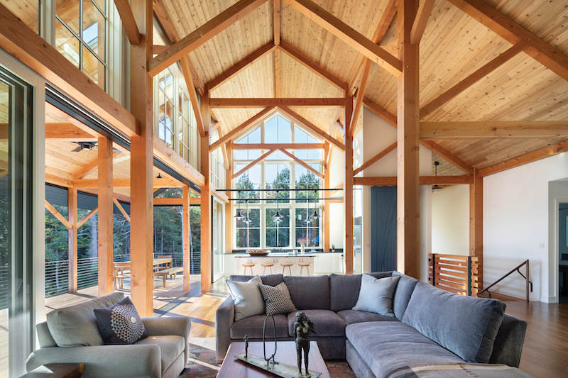 Two story living room with a kitchen in the background. There are a lot of windows and glass doors, a tongue and groove wood celling, and timber frame beams holding up the roof. 