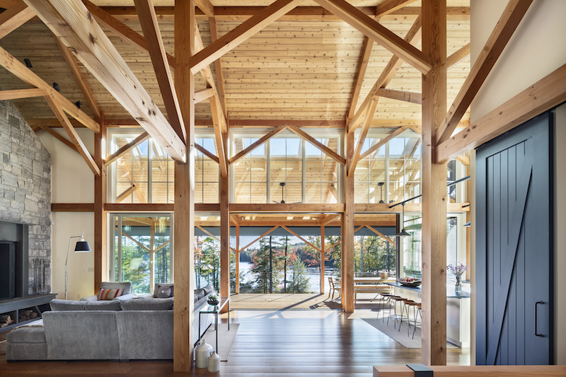 A large living area with a wood floor, lots of Marvin windows in the background and exposed timber framed beams holding the roof up. 