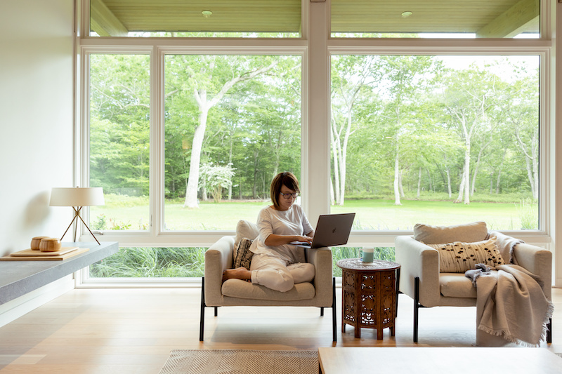A woman working on a laptop in a living room in front of a large bay of Marvin Ultimate casement and picture windows. 