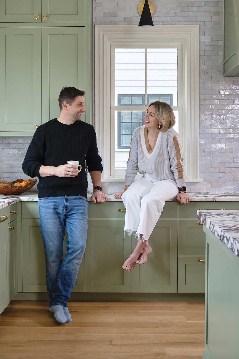 Sam Kachmar standing in his kitchen looking at his wife who is sitting on the marble countertop in front of a Marvin Ultimate double-hung window and sage green cabinets.