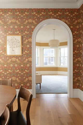 A rounded doorway from a dining room to a living space in an Italianate home, featuring floral wallpaper in reds, greens and golds.