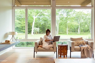 Woman sitting in a living room with laptop computer, Marvin Ultimate Casement Windows and Marvin Ultimate Picture Windows behind her looking out to a wooded lot.