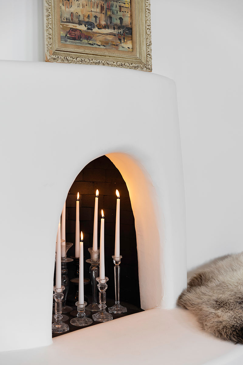 A Pueblo-style rounded white fireplace with lit tapered candles in glass candle holders inside.