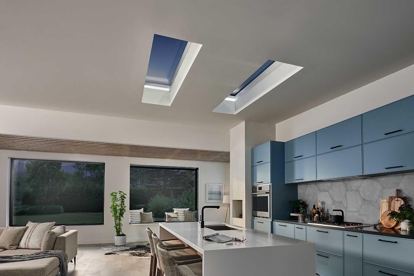 A modern kitchen at night with white countertops, blue cabinets, two Marvin Awaken Skylights and two Marvin Skycoves.