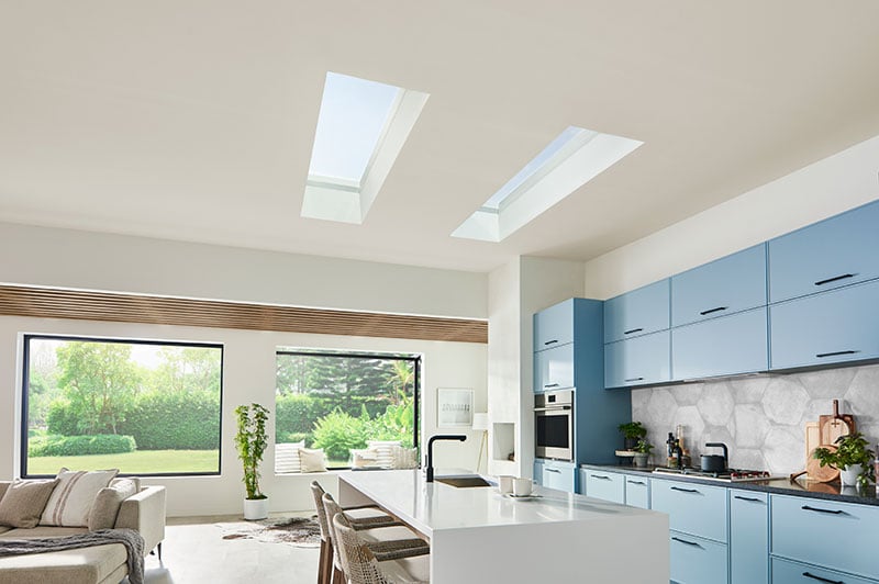 A modern kitchen with white countertops, blue cabinets, two Marvin Awaken Skylights and two Marvin Skycoves.