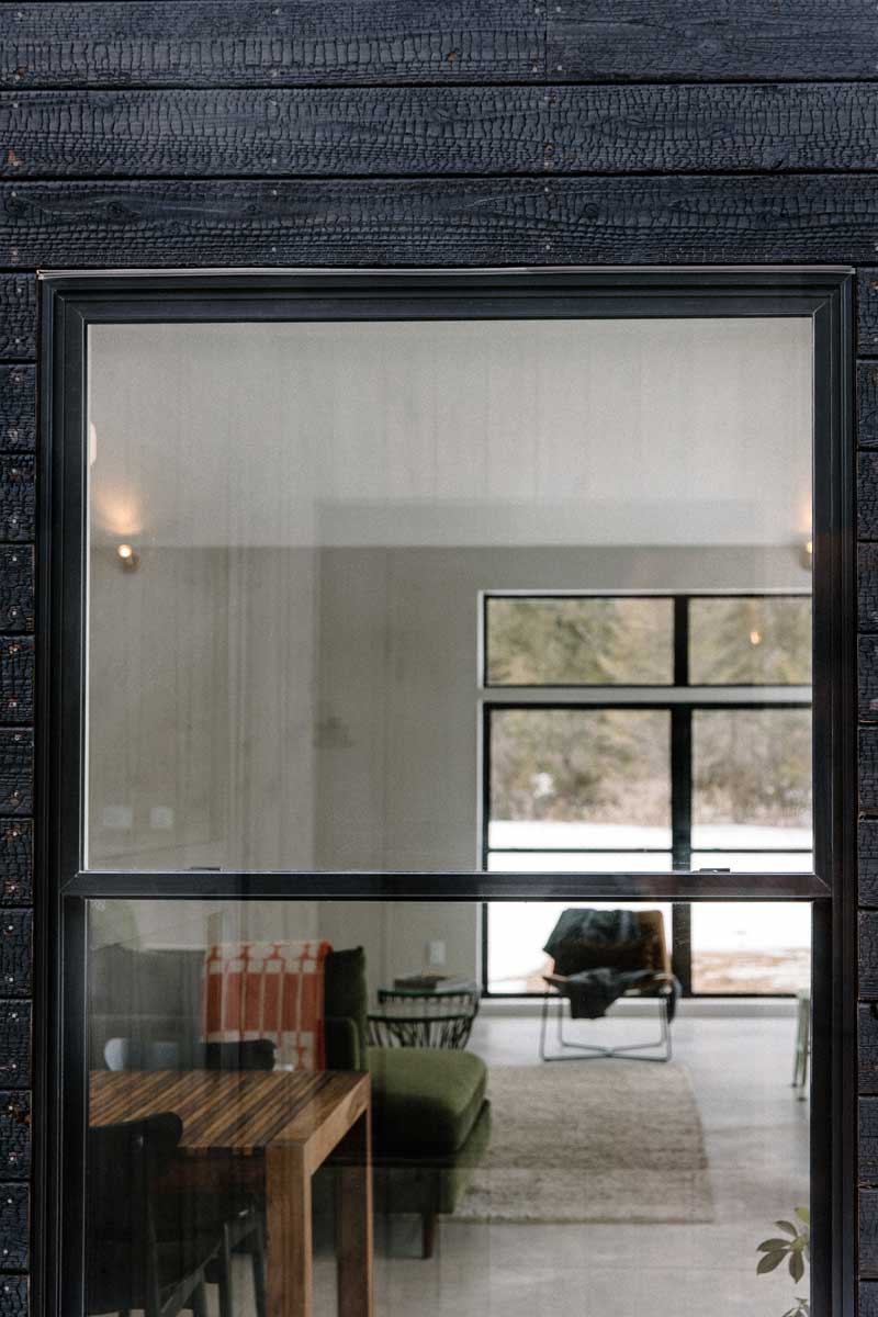 An exterior close up of a Marvin Essential double-hung window, framed in black siding, looking into the bright interior of a modern, minimalist living room. 