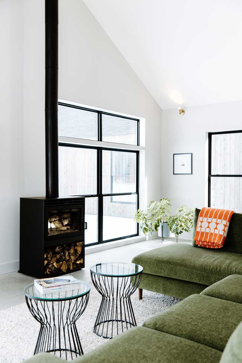 An angled view of the living room in Svart Hus showing a modern sage couch in front of a black wood burning stove, white walls, and the black framed Marvin Essential double-hung windows. 