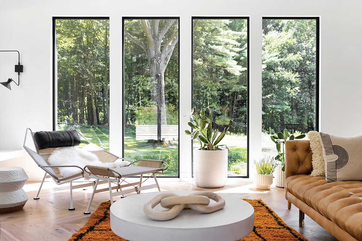 A modern seating area with a coffee table and plant with three large Marvin windows behind looking out to a lawn and large trees.