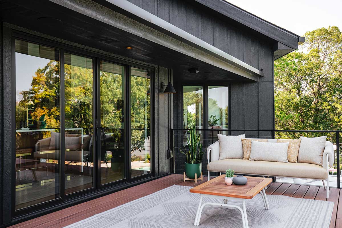 The back of a black home in Minnesota featuring a timber deck with mid-century style patio furniture and a Marvin Elevate Sliding glass door. You can see the reflection in the glass door that shows a tree-lined lake.