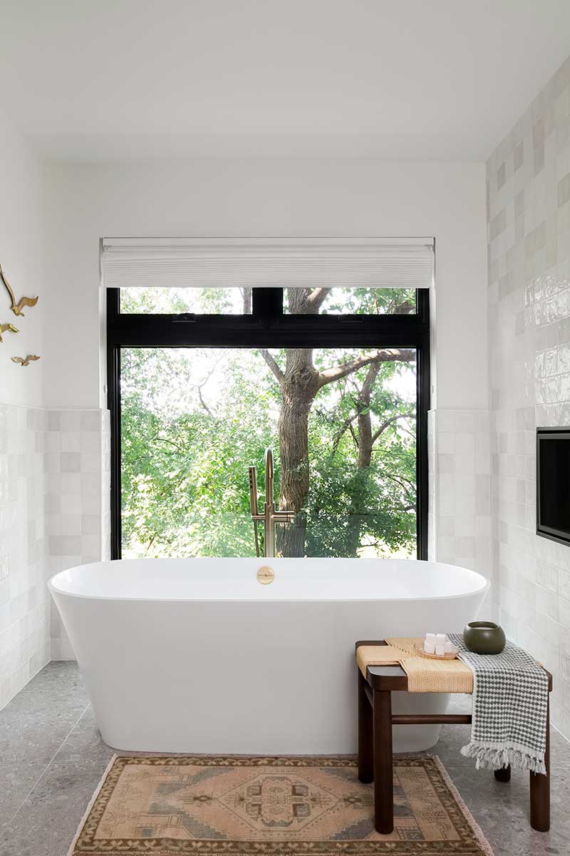 A large modern bathtub next to a footstool and rug with a large Marvin Essential window overlooking a tree-lined lake in Minnesota.