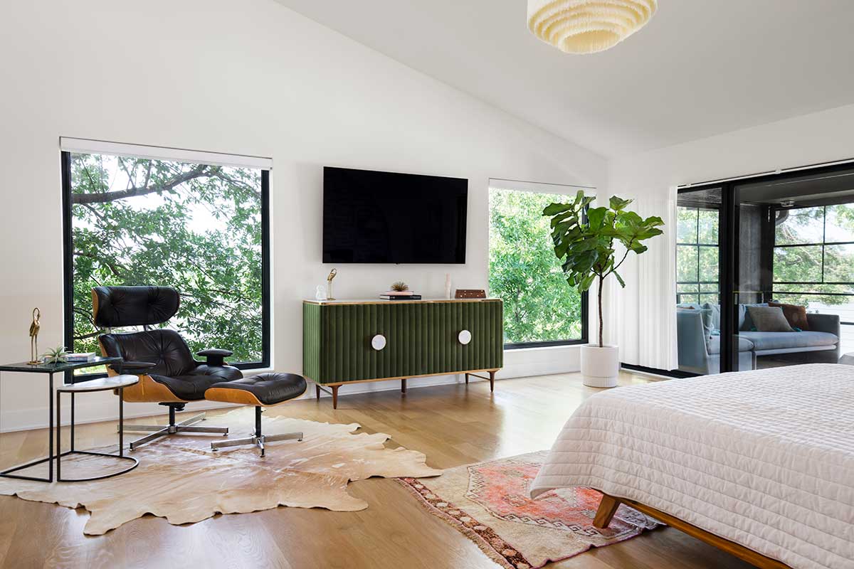 A mid-century styled bedroom with large Marvin Essential windows overlooking trees. A separate seating area can be seen through a Marvin Elevate sliding glass door.