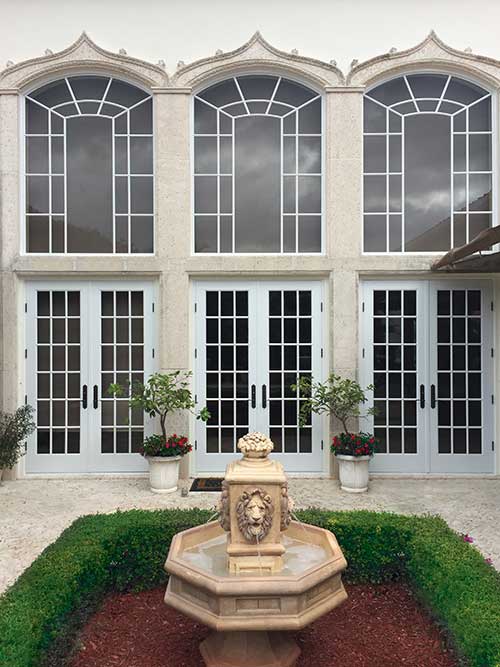 A courtyard with a lion head water fountain in the middle with three Marvin Coastline Outswing French doors and three Marvin Coastline Round Top Narrow Frame windows behind it. 