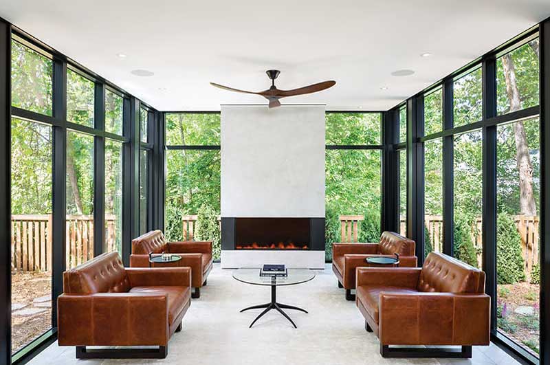A living room with a fireplace surrounded by floor-to-ceiling Marvin Modern Direct Glaze windows, four leather couches, a glass table, and a ceiling fan.