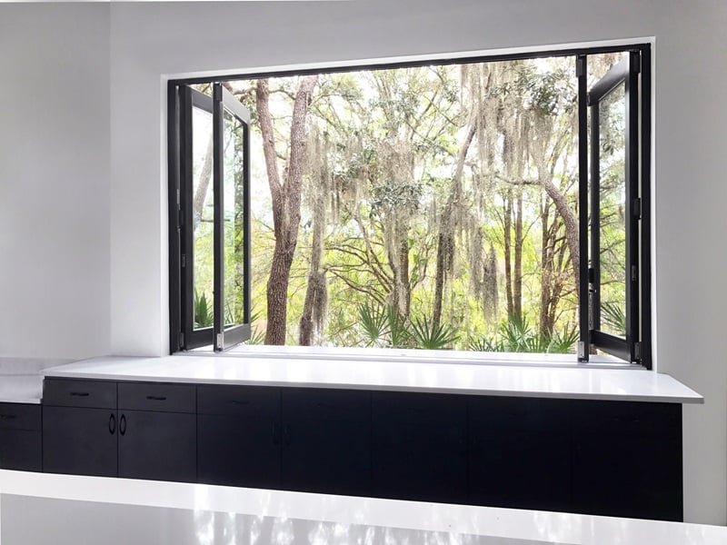 Marvin Bi-Fold Windows with a view of trees from inside a modern kitchen with white countertops and black cabinets.